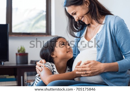 Lovely daughter touching mother's pregnant belly. Happy little girl feeling baby at mother tummy and awaiting the birth of her little brother. Pregnant mother relaxing on sofa with her cute daughter.