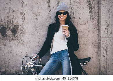 Lovely day. Beautiful young woman in sunglasses holding coffee cup and looking at camera with smile while standing against concrete wall outdoors  - Powered by Shutterstock