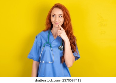 Lovely dreamy young red-haired doctor woman over yellow studio background keeps finger near lips looks aside copy space. - Powered by Shutterstock