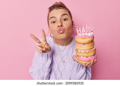 Lovely European woman blows mwah makes peace gesture shows victory sign holds pile of delicious doughnuts celebrates birthday wears sweater isolated over pink background. Celebration concept