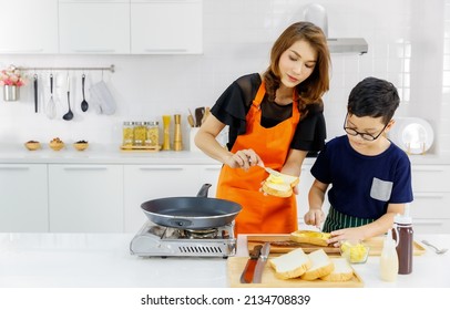 Lovely Family Education In Home Kitchen, Kid Learn To Cook Food And Mom On Orange Apron Standing  Beside Flying Pan And Gas Stove Happily Teaching Him To Put Butter On Flied Bread.