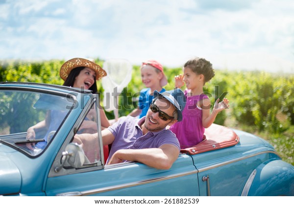 A lovely family is\
going on vacation in a convertible retro car. they drive on a\
country road on a sunny day