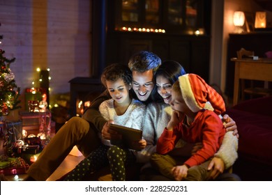 Lovely Family Sharing Digital Tablet Near The Wood Stove On A Winter Evening, Enjoying The Warm Christmas Atmosphere In Their Living Room, A Child Wears A Hat Of Santa Claus