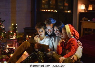 Lovely Family Sharing Digital Tablet Near The Wood Stove On A Winter Evening, Enjoying The Warm Christmas Atmosphere In Their Living Room, A Child Wears A Hat Of Santa Claus