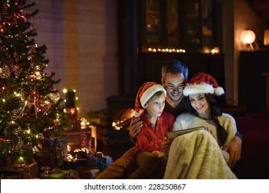 lovely family sharing digital tablet near the wood stove on a winter evening, enjoying the warm Christmas atmosphere in their living room, mother and child wearing a hat of Santa Claus - Powered by Shutterstock