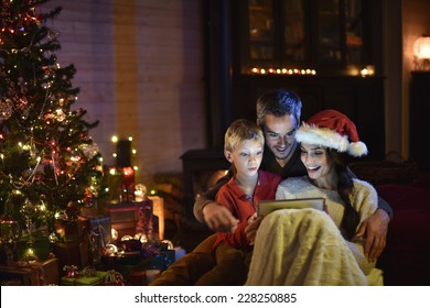 Lovely Family Sharing A Digital Tablet Near The Wood Stove On A Winter Evening, Enjoying The Warmth Of Christmas In Their Living Room, The Young Woman Wears A Hat Of Santa Claus