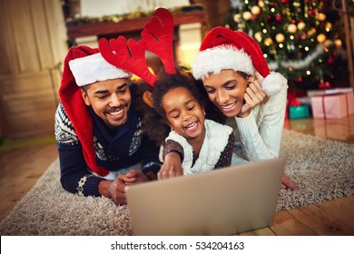 lovely family sharing laptop near the Christmas tree on a winter evening, enjoying the warm Christmas atmosphere in their living room and wearing a hat of Santa Claus