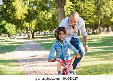 Lovely Father Teaching Son Riding Bike At Park. Happy Father Helping Excited Son To Ride A Bicycle At Park. Young Smiling Black Boy Wearing Bike Helmet While Learning To Ride Cycle With His Dad.