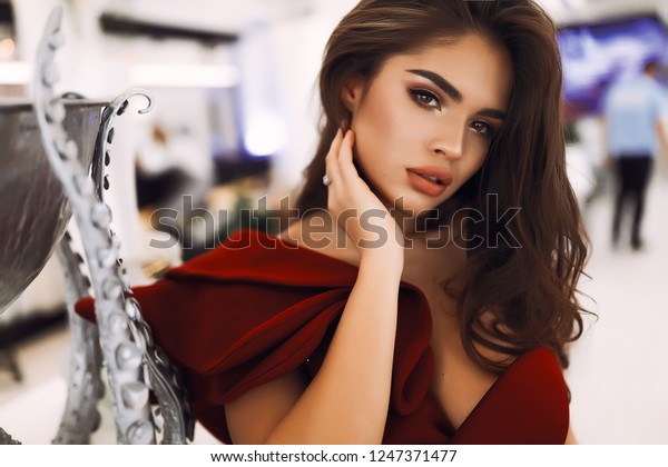 前を見る美しい女性 美しい目 大きな唇 ヌードの化粧 首に手を当て アクセサリーを持って 胸の上に黒い茶色の髪 銀色のデザイナーの花瓶の近くに立って赤いドレスを着て の写真素材 今すぐ編集