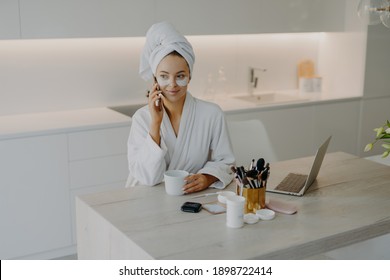 Lovely female model wears bathrobe and soft towel wrapped on head undergoes skin care procedures applies beauty patches under eyes has coffee break while talking with friend via cellular being at home - Powered by Shutterstock