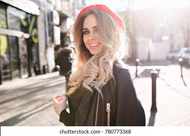 Lovely friendly woman with long blond hair wears red hat and leather jacket enjoys the sun and walks on street in sunlight with beautiful smile