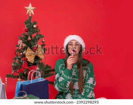 Lovely funny young woman sit near Christmas tree with shopping bags and present gift boxes. Plump lips fooling candy cane sweet. Isolated red background copy space. Happy new year concept.