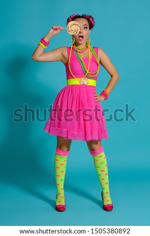 Lovely girl with a multi-colored braids hairstyle and bright make-up, posing in studio against a blue background, holding a lollipop in her hand.