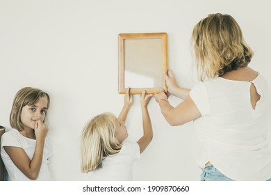 Lovely Girl Putting Frame On White Wall With Help Of Mom. Cute Daughter Hanging Blank Picture. Her Sister Standing Near And Looking Away. Family Decorating Room. Relocation And Moving Day Concept