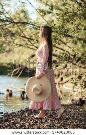 Lovely girl in a soft pink dress and a straw hat in her hands on the rocky shore of a beautiful river at sunset in summer. Image with selective focus