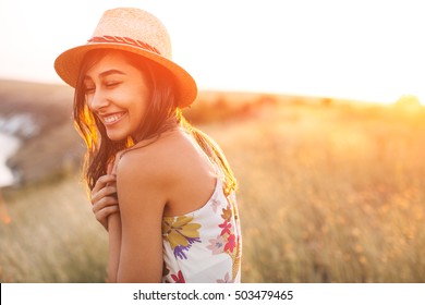 Lovely Girl In Stylish Hat And Summer Dress Smiling With Closed Eyes. Laughing Young Woman With Sun Light Flare Effect. Toned Image