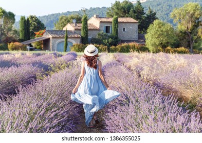Lovely girl walking by blooming lavender fields in Luberon area in Provence, France. Beautiful girl dressing straw hat and blue boho chic dress - traditional Provencal style.