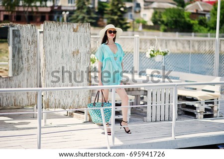 Lovely girl wearing transparent dress, hat and sunglasses walking on the quay by the lake with her bag.