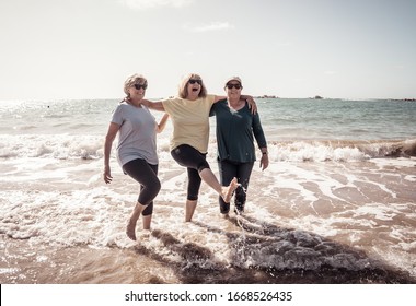 Lovely Group Of Senior Girl Friends On Their 60s Walking And Having Fun Splashing Water On Sea. Three Mature Healthy Retired Females Laughing And Enjoying Retirement And Outdoors Active Lifestyle.