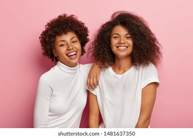 Lovely happy dark skinned women have fun together, enjoy spare time, smile happily, have natural curly hair, dressed casually, isolated on pink background. People, youth and emotions concept