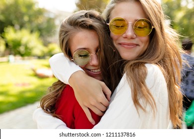 Lovely Happy Girls Walking Outside In Sunny Day. Beautiful Lovely Woman In Bright Glasses Hugging Her Friend And Closed Eyes With Great Smile, Best Friends, Sisters, Posivite Mood