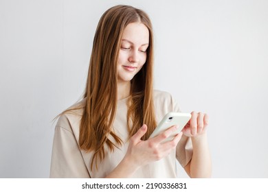 Lovely Happy Young Woman Holds Modern Cell Phone, Glad To Get Message From Boyfriend, Types Feedback, Checks Balance Online, Wears T Shirt Stands Over Gray Studio Wall