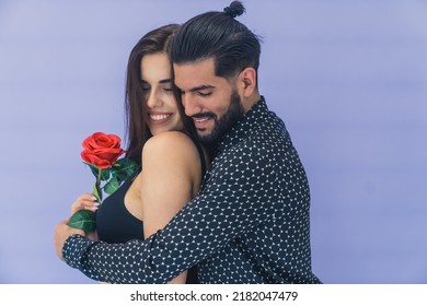 Lovely Heterosexual Couple Hugging In Studio Over Lavender Background. Handsome Bearded Girl Gives His Beautiful Girlfriend A Big, Red Rose. High Quality Photo