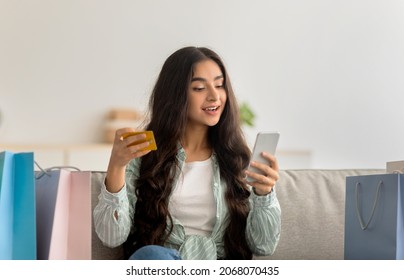 Lovely Indian Woman With Shopper Bags, Credit Card And Smartphone Shopping Online From Home, Happy Over Huge Sale In Web Store. Young Eastern Lady Buying Goods On Internet