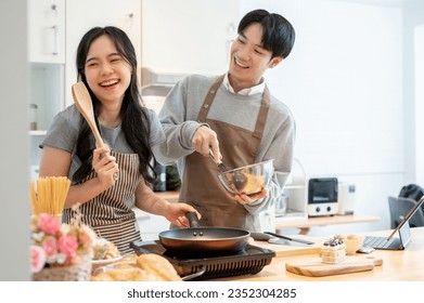 A lovely and joyful young Asian couple is laughing, playing, enjoying baking pastry, or making pancakes in the kitchen together. cooking dates at home, leisure, family activity - Powered by Shutterstock