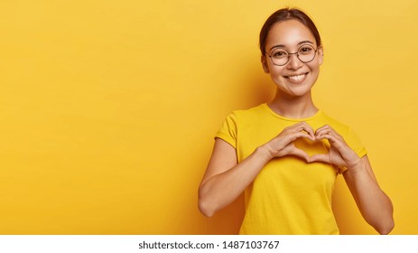 Lovely korean woman with happy smile, shapes heart with both hands, expresses love to you, wears round spectacles and yellow t shirt, says be my valentine flirts with boyfriend poses indoor copy space