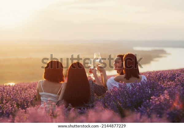 Lovely ladies drinking wine in lavender
violet field at sunset. Summer happy mood. Girlfriends relaxing on
summer sunset with river on the
background.