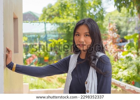 A lovely lady in a cardigan and navy blue long sleeves top is looking directly at the camera with a straight face while her hand is leaning on a wall. Plants and trees in the background.