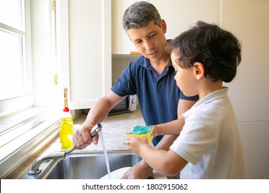 Lovely little boy helping dad, holding wet sponge and washing plates in kitchen sink. Happy handsome father holding faucet with water and looking at son. Housekeeping and family concept - Powered by Shutterstock