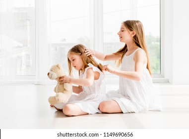 Lovely little girl brushing hair of her younger sister while sitting on the floor. Little child girl playing with bear toy while her older sister combing her hair