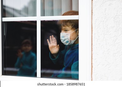 Encantador niño pequeño de la escuela por una ventana con máscara médica en casa o en la escuela durante la cuarentena del coronavirus pandémico. Un niño solitario y disgustado que mira a su mejor amigo y wafes.
