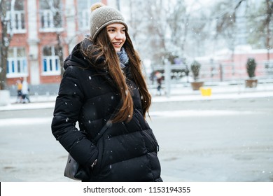 the lovely long-haired girl in a warm black jacket and hat walks through the winter city, smiling