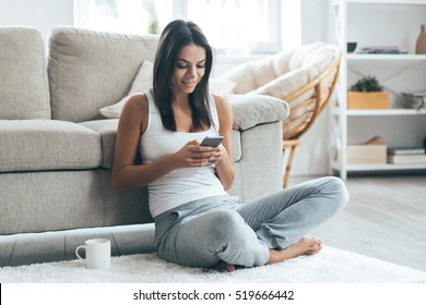 Lovely message from him. Attractive young woman looking at her smart phone and smiling while sitting on the carpet at home 
