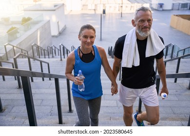 Lovely Middle Aged Couple Of Sportive Man And Woman Looking Happy, Holding Hands While Walking Up The Stairs After Exercising Together In The City