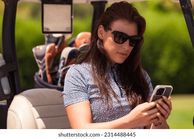 Lovely middle aged woman golfer in sunglasses sitting on golf cart in golf club and using mobile phone, looking at cellphone. Healthy sports lifestyle and leisure activity concept. Copy ad text space - Powered by Shutterstock