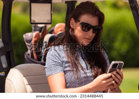 Lovely middle aged woman golfer in sunglasses sitting on golf cart in golf club and using mobile phone, looking at cellphone. Healthy sports lifestyle and leisure activity concept. Copy ad text space