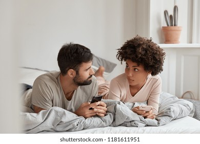 Lovely mixed race couple have pleasant talk, use mobile phones in bed at morning, look seriously at each other, enjoy coziness at bedroom, free wifi connection at home. Contemporary technology - Powered by Shutterstock