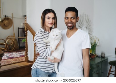 Lovely mixed-races young happy joyful couple male and female smiling in good mood resting at home in living room with pet animal holding cat in hands, pet lover concept