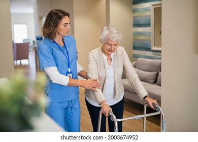 Lovely Nurse Helping Old Woman To Walk At Nursing Home With Walker. Young Nurse Helping Senior Patient Using A Walking Frame To Walk In Hospital Corridor. Caregiver And Disabled Lady In Care Facility.