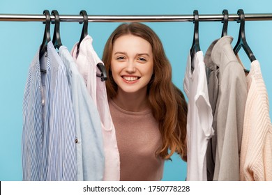 Lovely Positive Woman, Fashion Stylist Looking Out Of Clothes Hang On Shelf In Designer Store, Smiling Playfully And Hiding In Wardrobe With Trendy Outfit On Rack. Indoor Studio Shot, Isolated