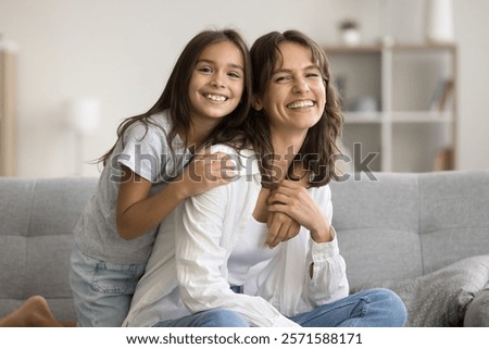 Lovely preschooler daughter embracing her young mommy, sitting together on couch in living room, laughing, looking at camera, resting at home. Happy Mother Day celebration, family ties, motherhood