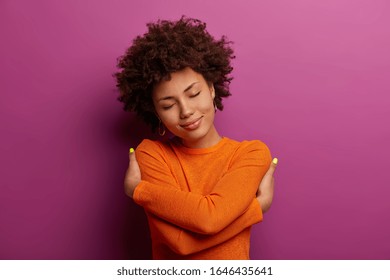 Lovely pretty woman embraces herself, feels good, comfortable and fullfilled, has high self esteem, tilts head and closes eyes, being egoisitc person, wears orange jumper isolated on purple background - Shutterstock ID 1646435641