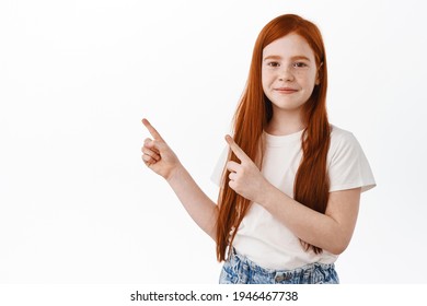 Lovely Red Head Kid Girl With Freckles, Ginger Child Pointing Fingers At Upper Left Corner, Smile Shy, Standing Over White Background In Casual T-shirt