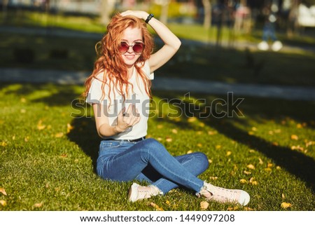 A lovely red-haired girl, warmed by the rays of the sun, is sitting on a lawn and posing for the camera. The girl is wearing a T-shirt with jeans, glasses on her face, and a modern gadget on her arm.