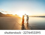 Lovely, romantic couple walking on sunset beach, enjoying evening light, relaxing on tropical summer vacation. Honeymoon. Love. Back view. Woman wearing orange maxi dress.
