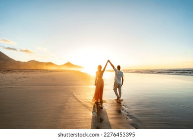 Lovely, romantic couple walking on sunset beach, enjoying evening light, relaxing on tropical summer vacation. Honeymoon. Love. Back view. Woman wearing orange maxi dress.
